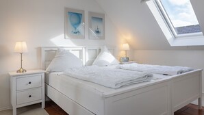 Un dormitorio con tragaluces, una cama con ropa de cama blanca, dos mesitas de noche con lámparas y dos cuadros colgados en la pared.