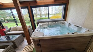 Een kuip met warm water, een houten buitenkant en een glazen omheining, omringd door rieten meubels en een gebied met kunstgras.
