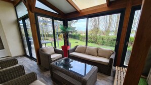 Een zonnige kamer met rieten meubels, een glazen tafel en uitzicht op een tuin door grote ramen.