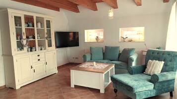Een woonkamer met een houten plafond, een televisie, een kast met glazen deuren, een blauwe bank en een salontafel.
