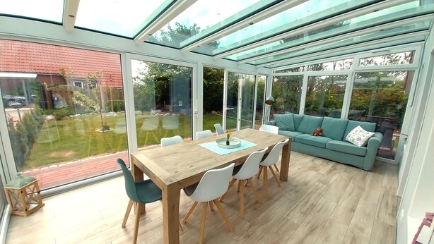 A conservatory with a wooden table and chairs, a teal sofa, and a view of the garden and neighboring buildings.