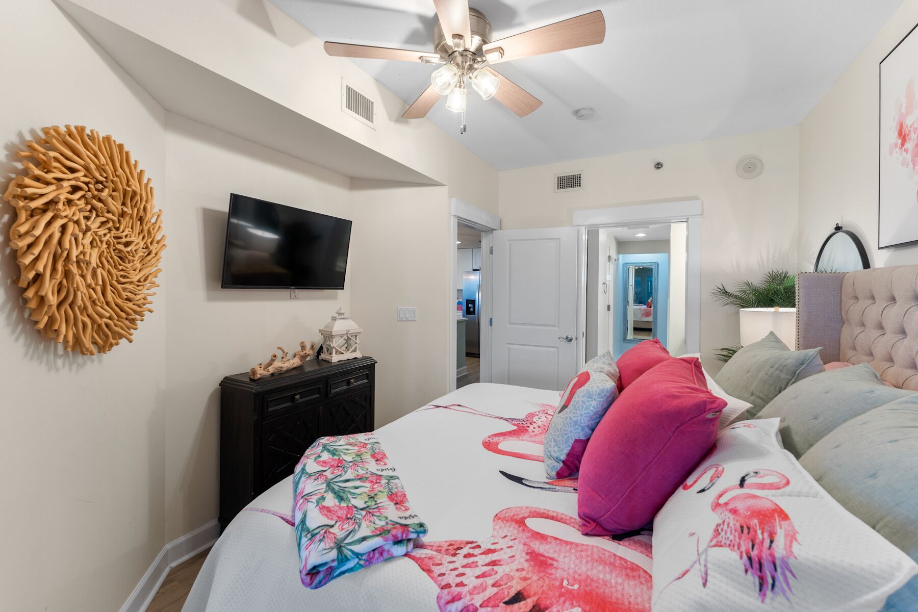 A bedroom with a bed, a ceiling fan, a TV, a dresser, and a door leading to another room.