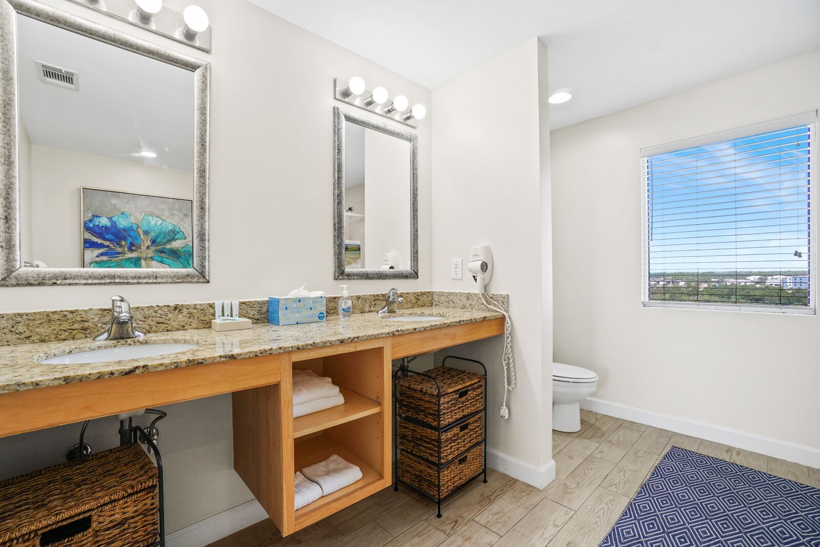 A bathroom with a granite countertop, two mirrors with lights, a sink, a toilet, and a window with a view.