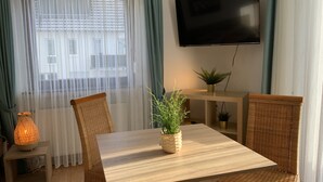 Una habitación con una mesa de madera, sillas de mimbre, un televisor fijado a la pared y una ventana con cortinas traslúcidas.