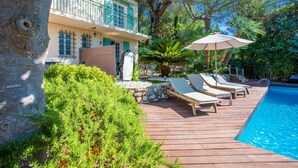 A wooden deck with lounge chairs and an umbrella, a swimming pool, and a building with a balcony. - VILLA JUSTINE VI4239 By Riviera Holiday Homes (Mandelieu-la-Napoule)