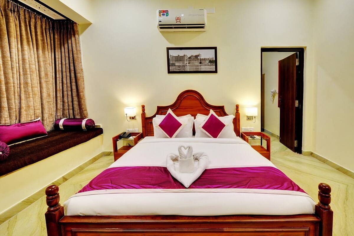 A hotel room with a large bed, a wooden headboard, a window with curtains, an air conditioning unit, a framed picture on the wall, and a door.
