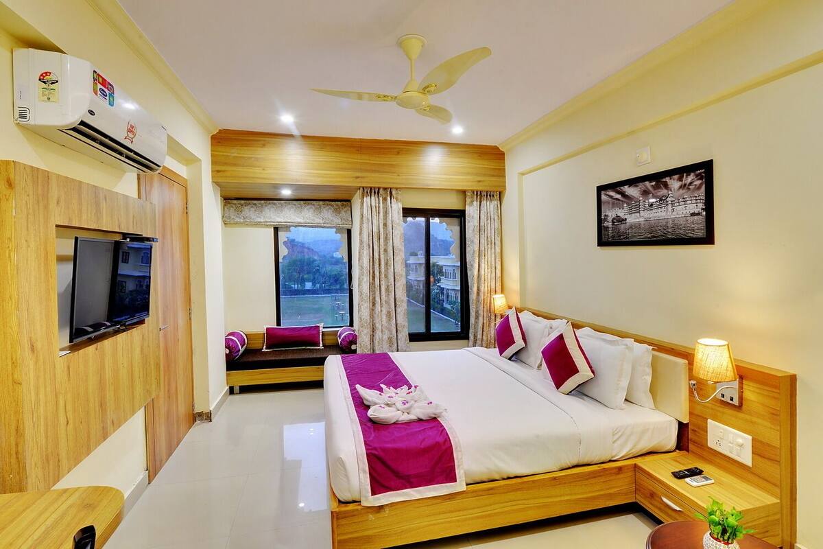 A hotel room with a bed, bedside table, lamp, and a view of a building through the window.