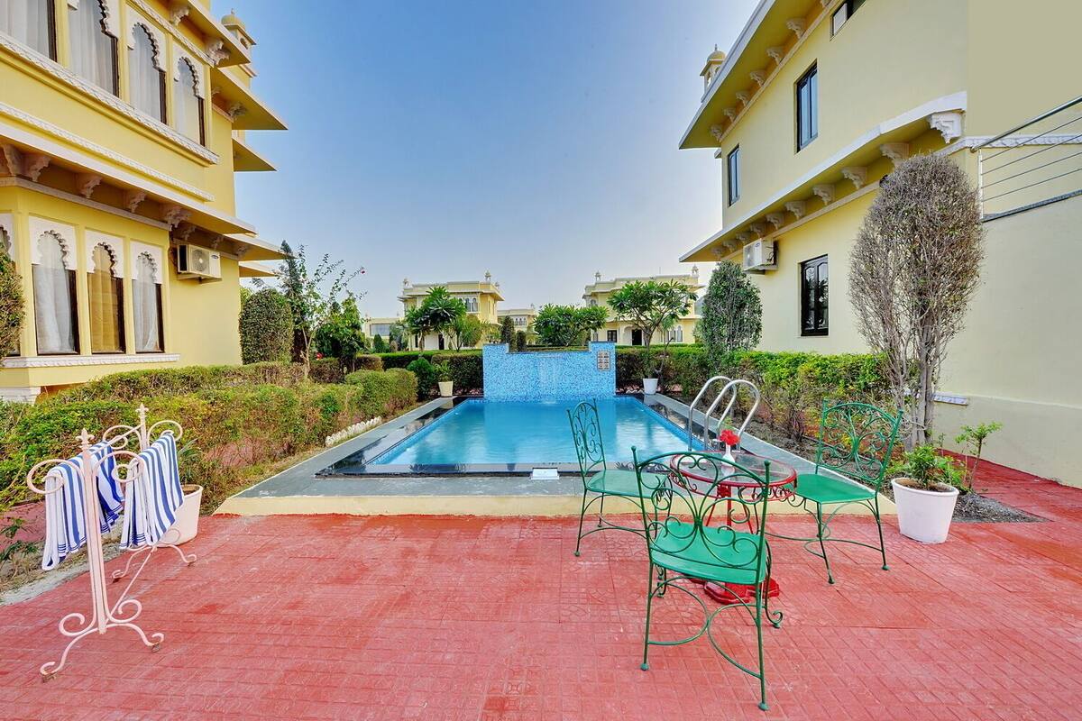 A swimming pool with a red-tiled deck, surrounded by greenery and a building with yellow walls and ornate windows.