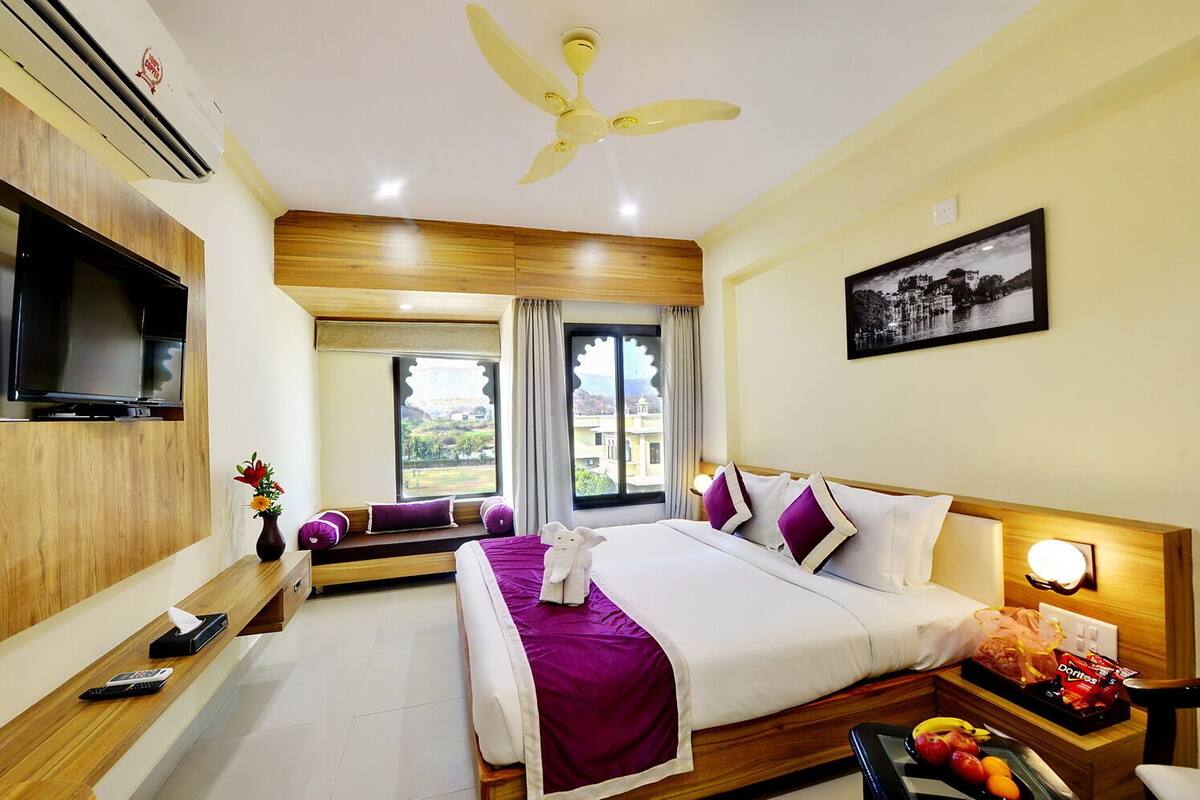 A hotel room with a bed, a television, a window with a view, a ceiling fan, and a nightstand with snacks.