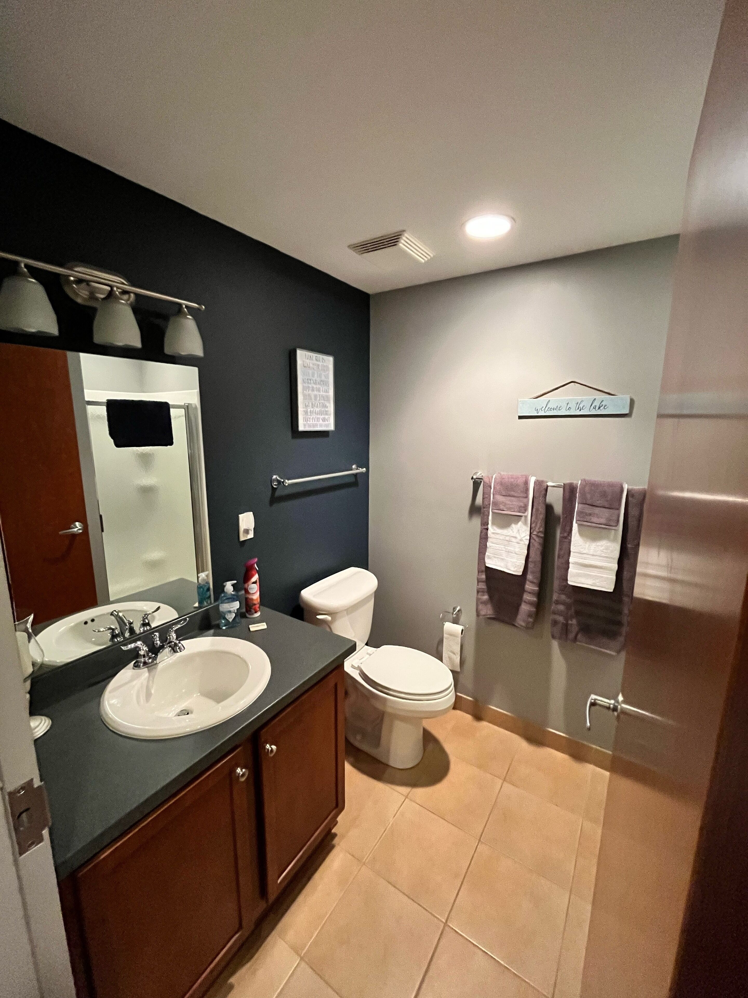 Une salle de bain avec un double lavabo, des toilettes et un espace douche. On y trouve un porte-serviettes avec des serviettes et une pancarte sur laquelle est écrit : « Bienvenue au bord du lac ».