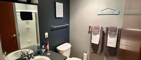 Une salle de bain avec un double lavabo, des toilettes et un espace douche. On y trouve un porte-serviettes avec des serviettes et une pancarte sur laquelle est écrit : « Bienvenue au bord du lac ».