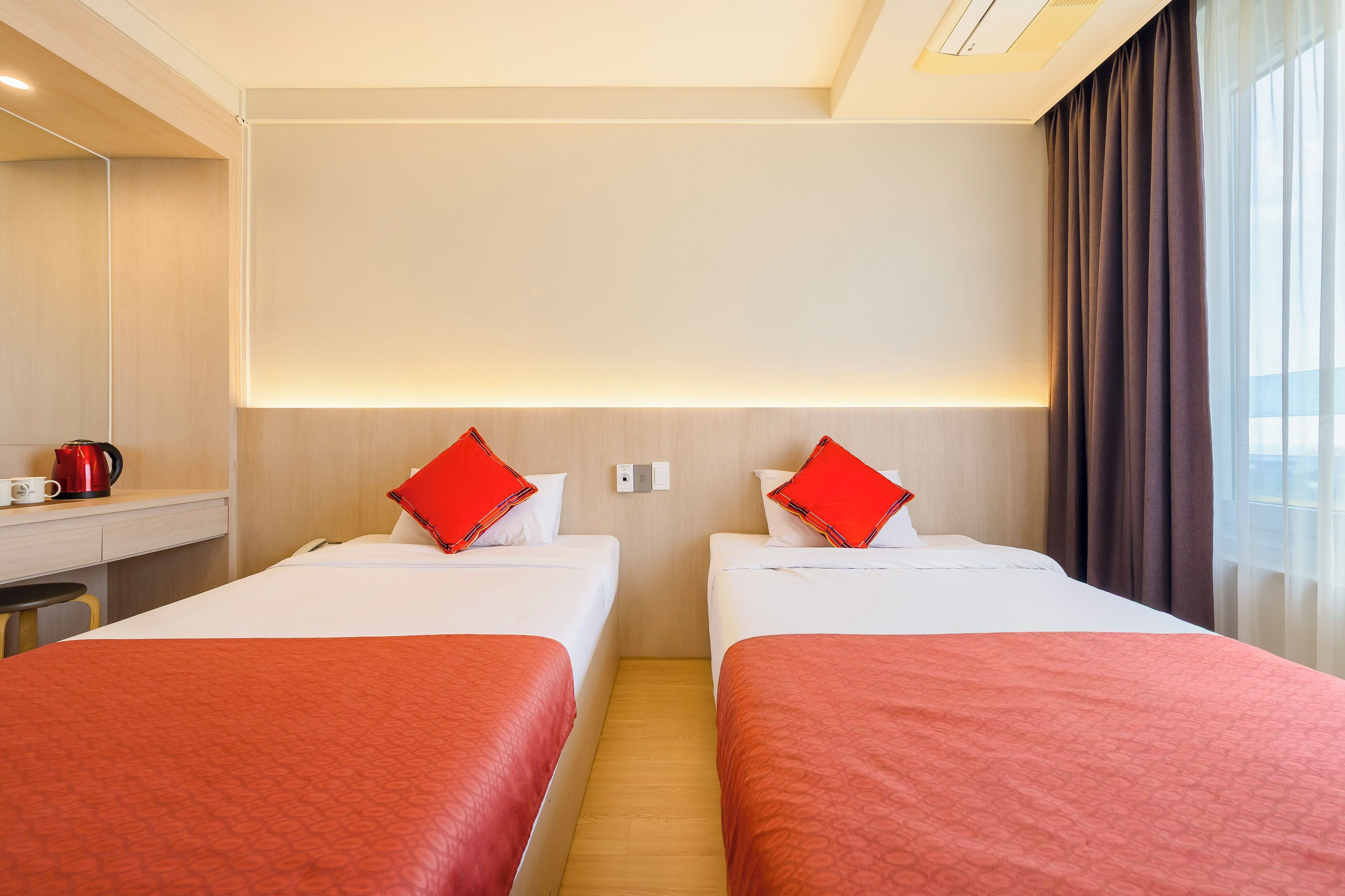 Quarto de hotel com duas camas, cada uma com colcha e almofadas vermelhas, uma escrivaninha com chaleira vermelha e uma janela grande com cortinas.