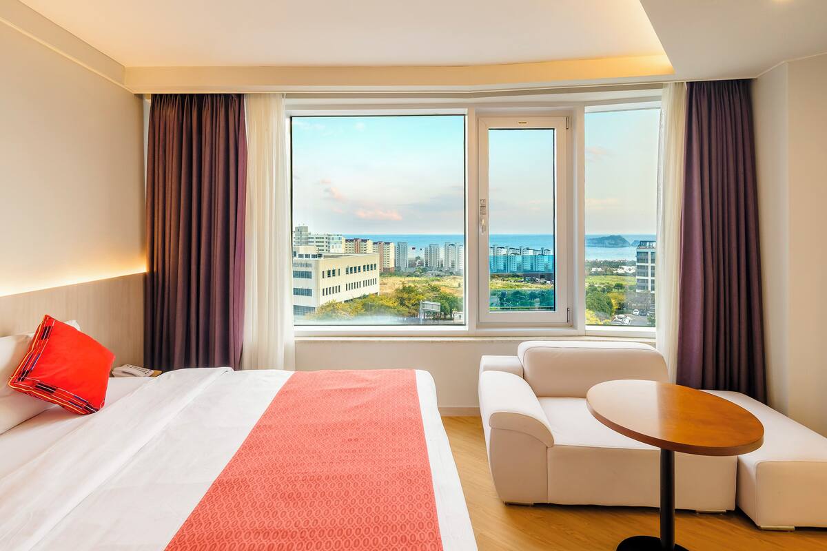 A hotel room with a large bed, a small sofa, a round table, and a view of the city through large windows.