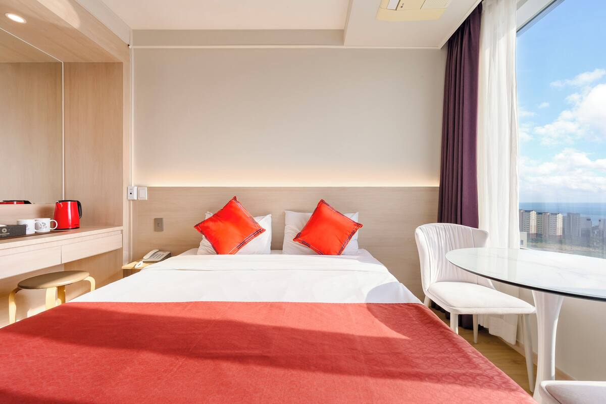 A hotel room with a bed, a desk with a telephone, a coffee maker, a glass table, and a chair.