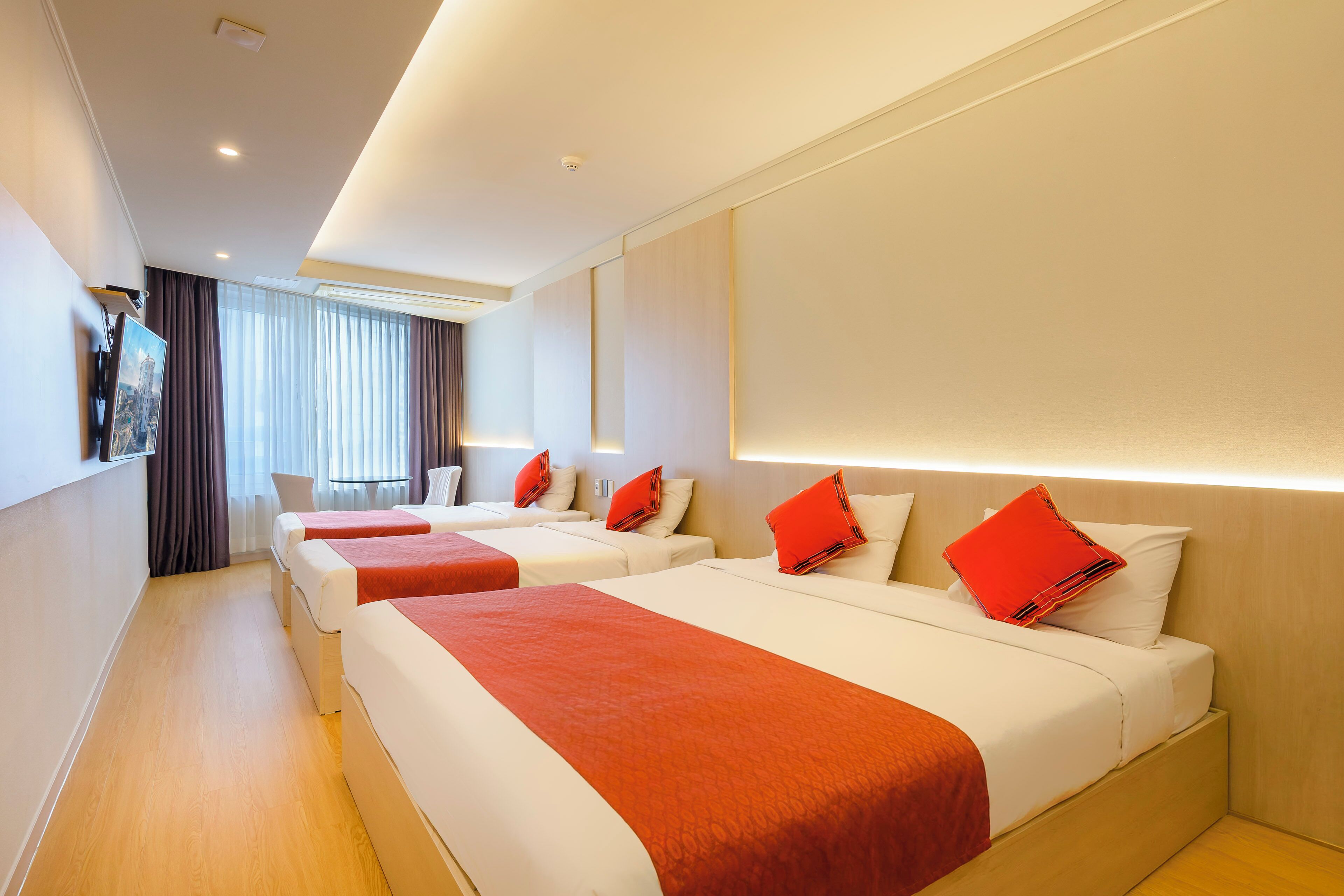 A hotel room with three beds, each with red and white bedding, and a large window with curtains.