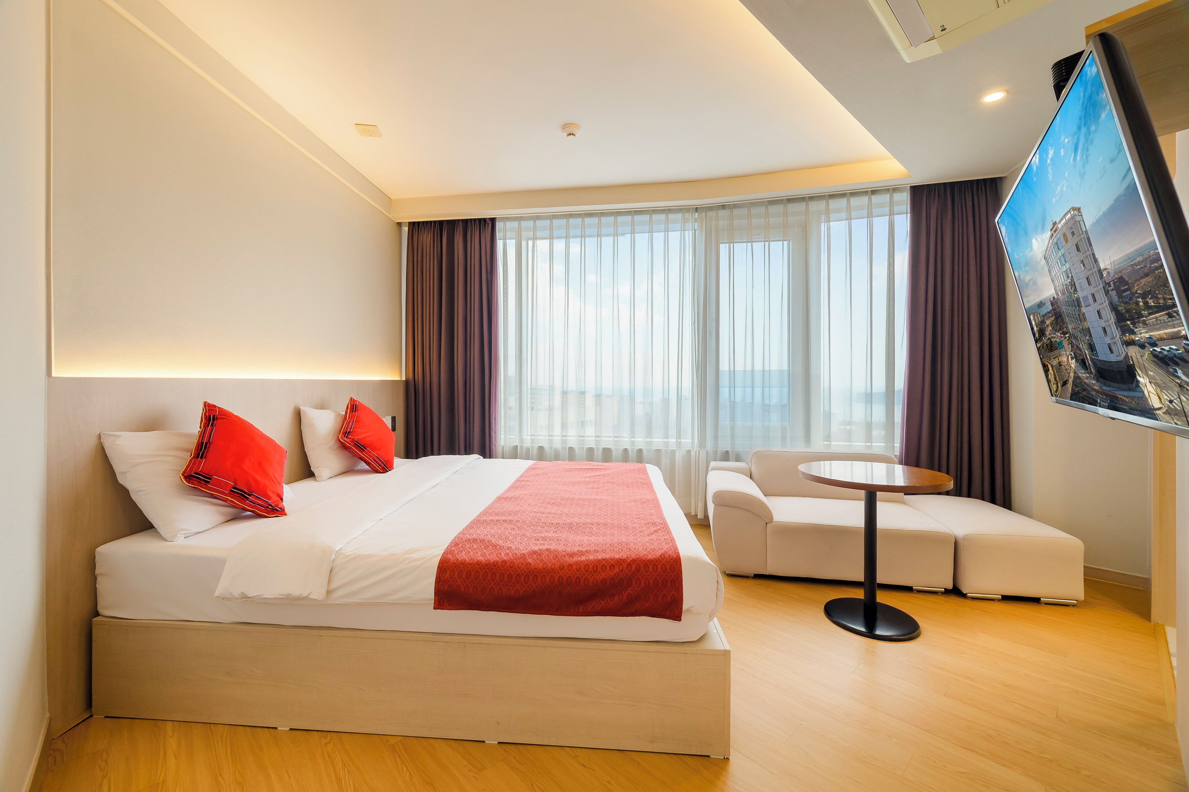 A modern hotel room with a large bed, a small sofa, a round table, and a TV on the wall.