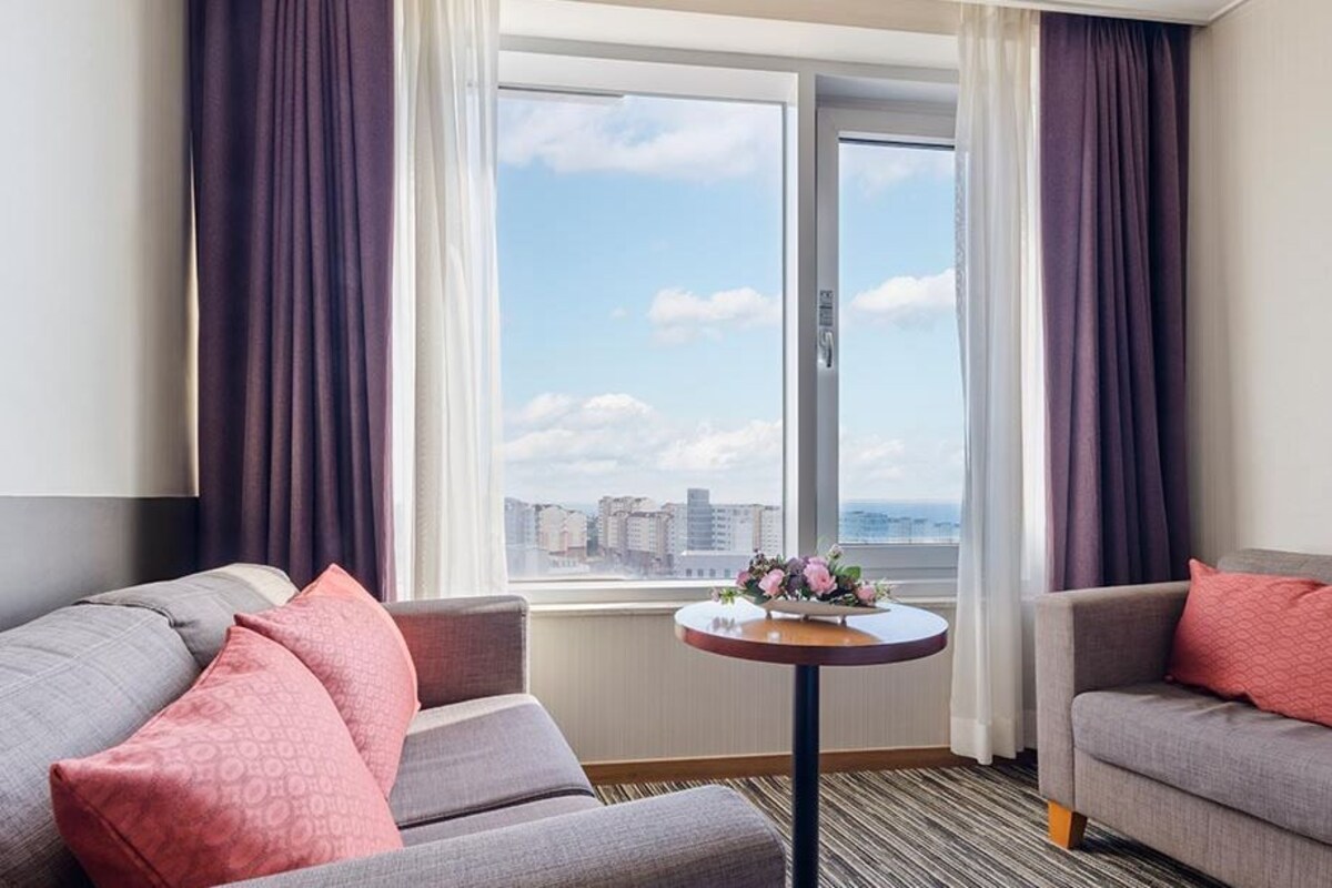 Habitación de hotel con dos sofás grises, una mesita redonda con un jarrón de flores y un ventanal con vistas a la ciudad.