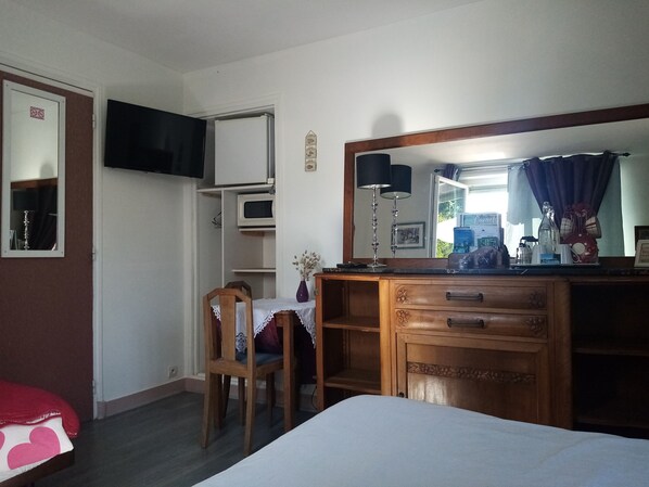 A bedroom with a bed, a wooden dresser, a desk with a chair, a television, and a microwave.