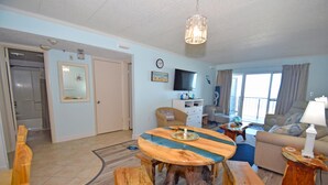 A living room with a round wooden coffee table, a sofa, and a television. There is a door leading to a bathroom and a window with curtains. - Marigot Beach 303-Oceanfront 101st St, Free WiFi, Pool, Elev, W/D, AC (Ocean City)