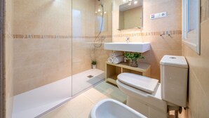 Une salle de bain moderne avec une douche à parois de verre, un lavabo blanc, des toilettes et une petite étagère avec des serviettes et une plante.