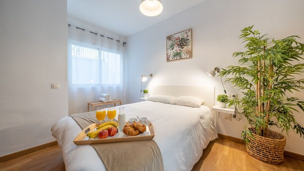 Une chambre d’hôtel avec un lit, une table de chevet avec une lampe, une fenêtre avec des rideaux légers, une œuvre d’art accrochée au mur et une grande plante en pot.