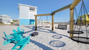 A beachside area with turquoise Adirondack chairs, a swing set, and a building with a blue and white facade. - Family Friendly with Gameroom! Outdoor swings and more! (Navarre)