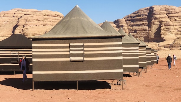 A row of simple, striped tents with conical roofs, set against a desert landscape with rocky formations.