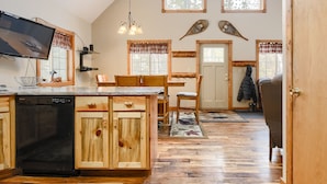 A kitchen with wooden cabinets, a dishwasher, a dining table with chairs, a TV, and a large window with a view of trees. - 3-Level Spacious Cabin w/ Pool Table, Foosball, Close to Terry Peak & Skiing (Lead)