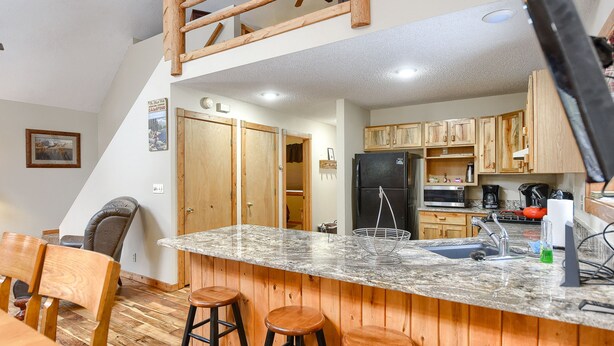 A kitchen with wooden cabinets, a granite countertop, and a built-in sink. There are three stools and a chair in the dining area. - 3-Level Spacious Cabin w/ Pool Table, Foosball, Close to Terry Peak & Skiing (Lead)