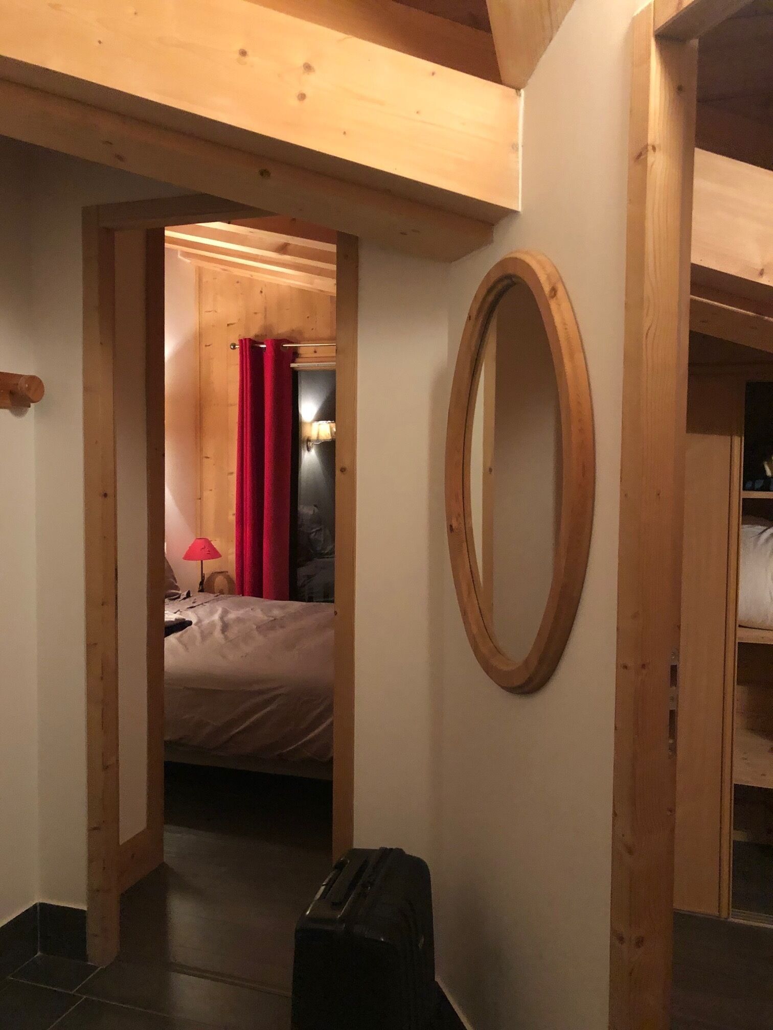 A hotel room with a wooden ceiling, a bed with a red curtain, a small table with a lamp, and a suitcase on the floor.