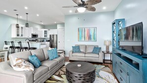 A modern living room with a sofa, coffee table, and TV stand. There is a kitchen in the background with white cabinets and a dining area with chairs. - Pet Friendly Gulf View Townhome | Sleeps 10 | Game Room (Pensacola Beach)