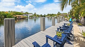A waterfront dock with wooden posts, a row of blue chairs, and a view of a canal with houses and greenery in the background.