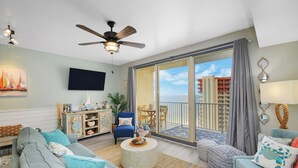 A spacious living room with a large sofa, a round coffee table, and a blue armchair. A ceiling fan and a flat-screen TV are mounted on the wall. The room has a large sliding glass door leading to a balcony with a view of buildings. - Beach Front Free round of Golf & Activities Daily! (Panama City Beach)