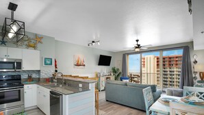 A modern kitchen with a central island, a dining area with a table and chairs, a living room with a sofa, and a balcony with a view of buildings. - Beach Front Free round of Golf & Activities Daily! (Panama City Beach)