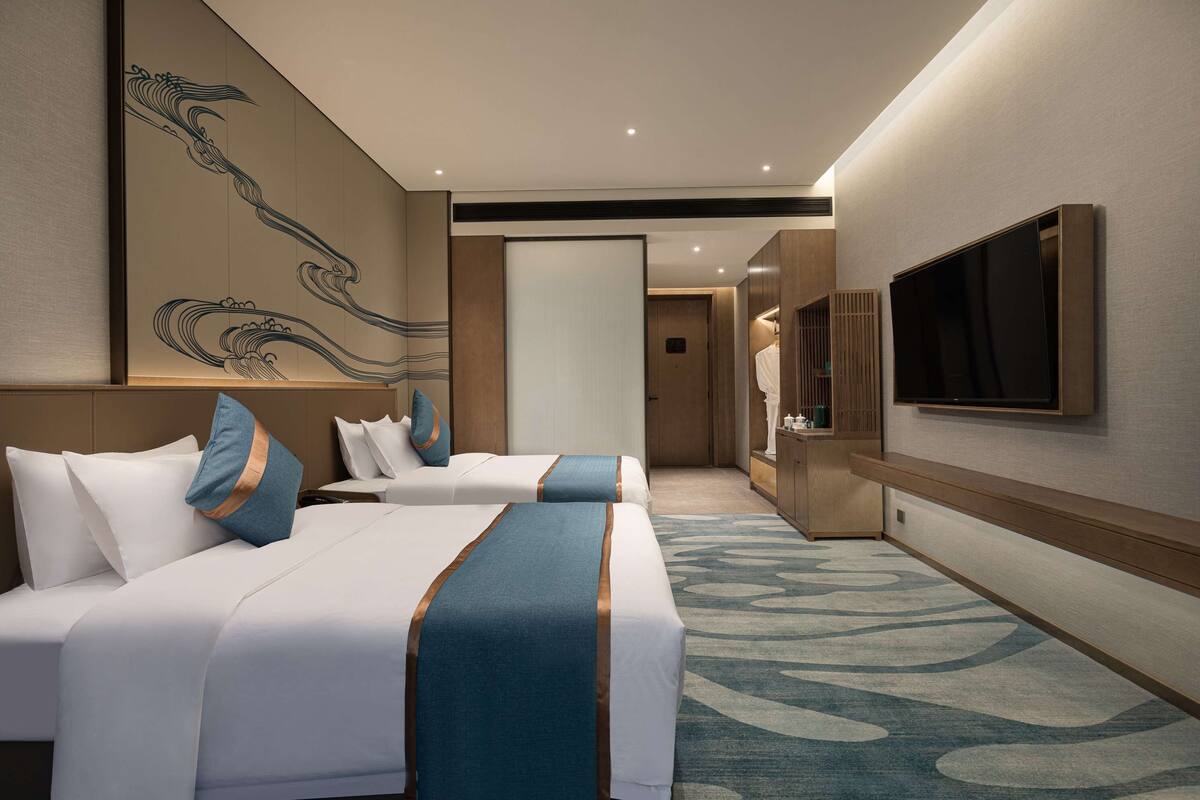 A hotel room with two beds, a flat-screen TV mounted on the wall, and a large window.