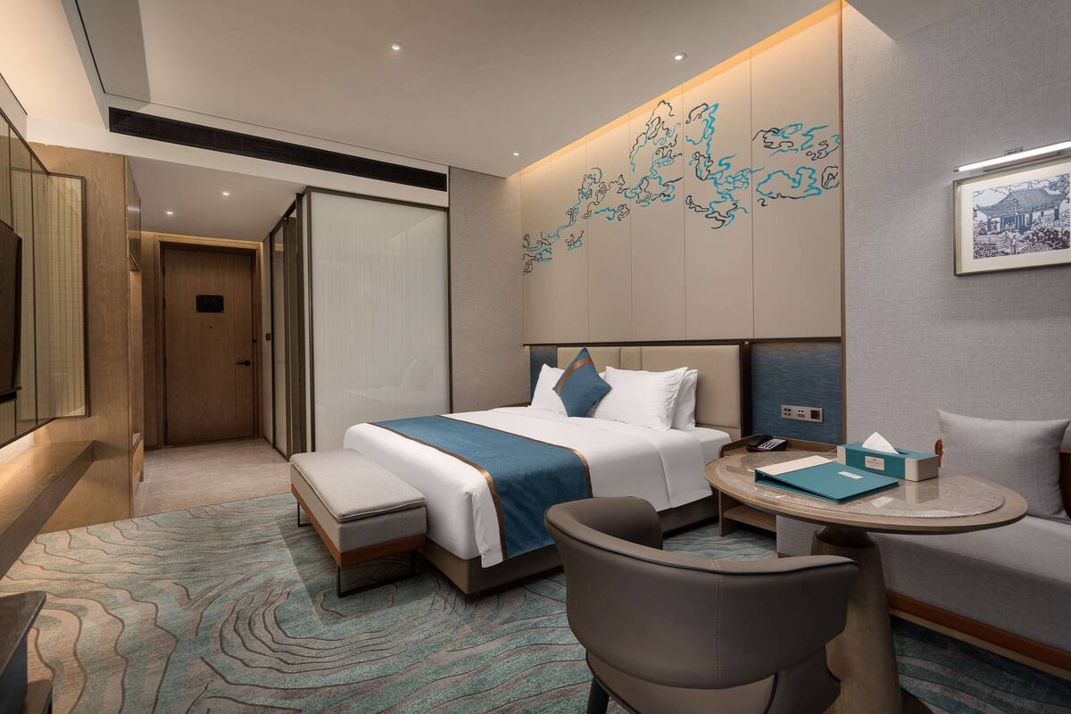 A modern hotel room with a neatly made bed, a small round table with a chair, a sofa, and a large artwork on the wall.