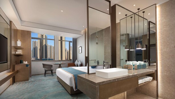 Habitación de hotel moderna con una cama grande, zona de estar, baño con espejo grande y lavamanos, y vistas a edificios altos.