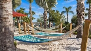 A hammock on a sandy beach with palm trees and a red umbrella in the background. - Upgraded! Stunning Pool, Stylish Decor! Kona Cove Retreat, Prime Disney! (Davenport)