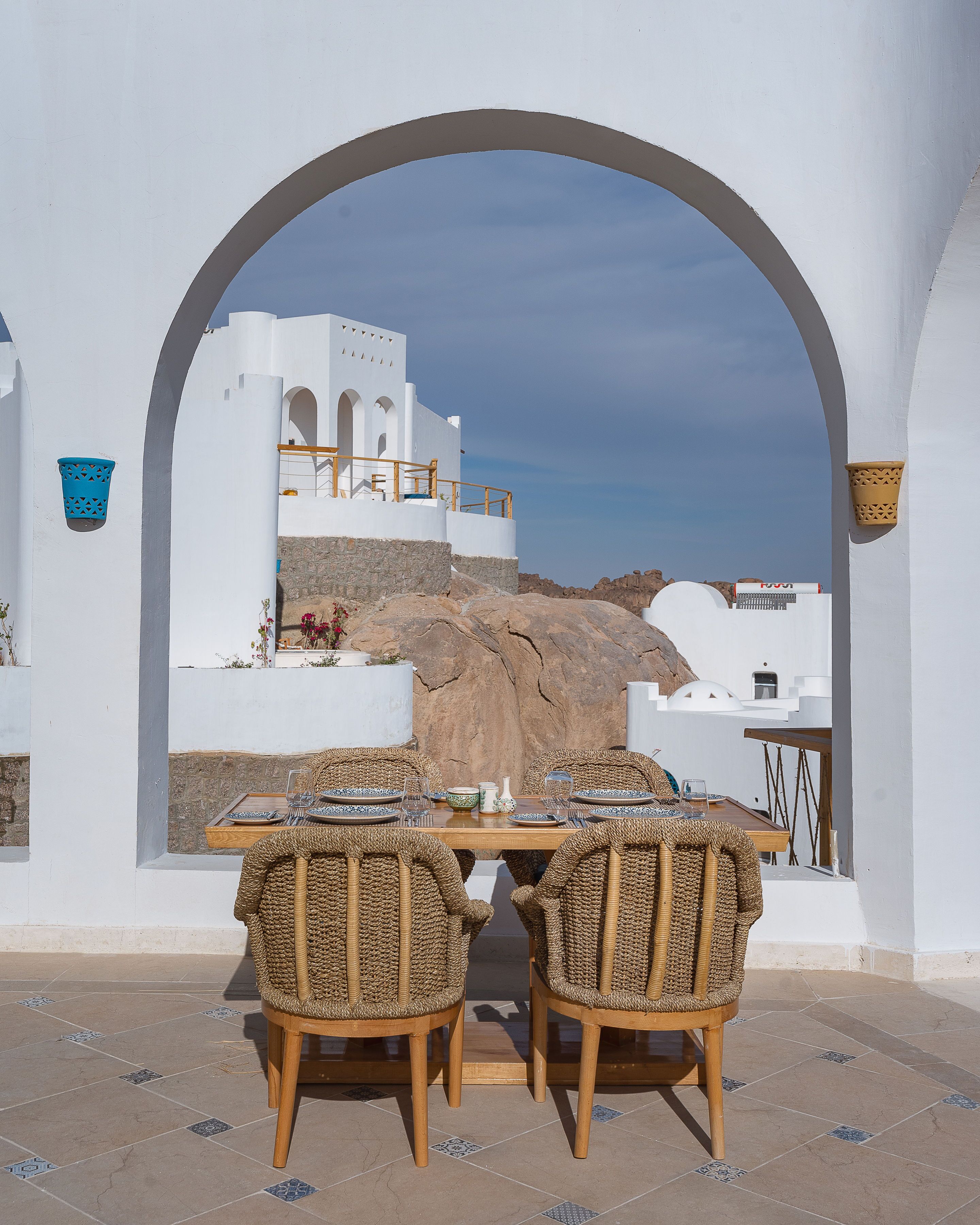 A dining table set with wicker chairs, white walls, and a view of a white building with a stone base.