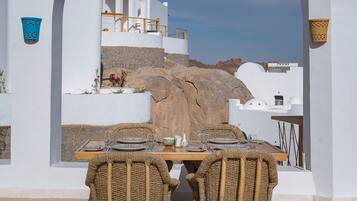 A dining table set with wicker chairs, white walls, and a view of a white building with a stone base.