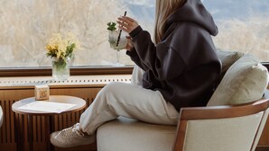 Eine Person sitzt am Fenster mit Blick auf Berge, an einem kleinen Tisch steht eine Vase mit Blumen und ein Getränk.