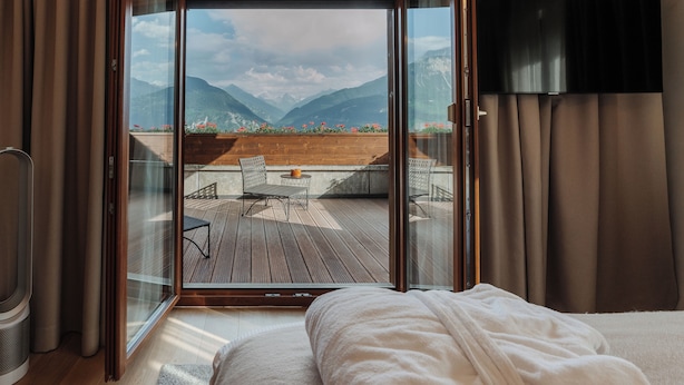 Ein Schlafzimmer mit einer großen Schiebeglasdoor, die auf einen Balkon mit Holzterrasse, einem Stuhl und einem Tisch führt.