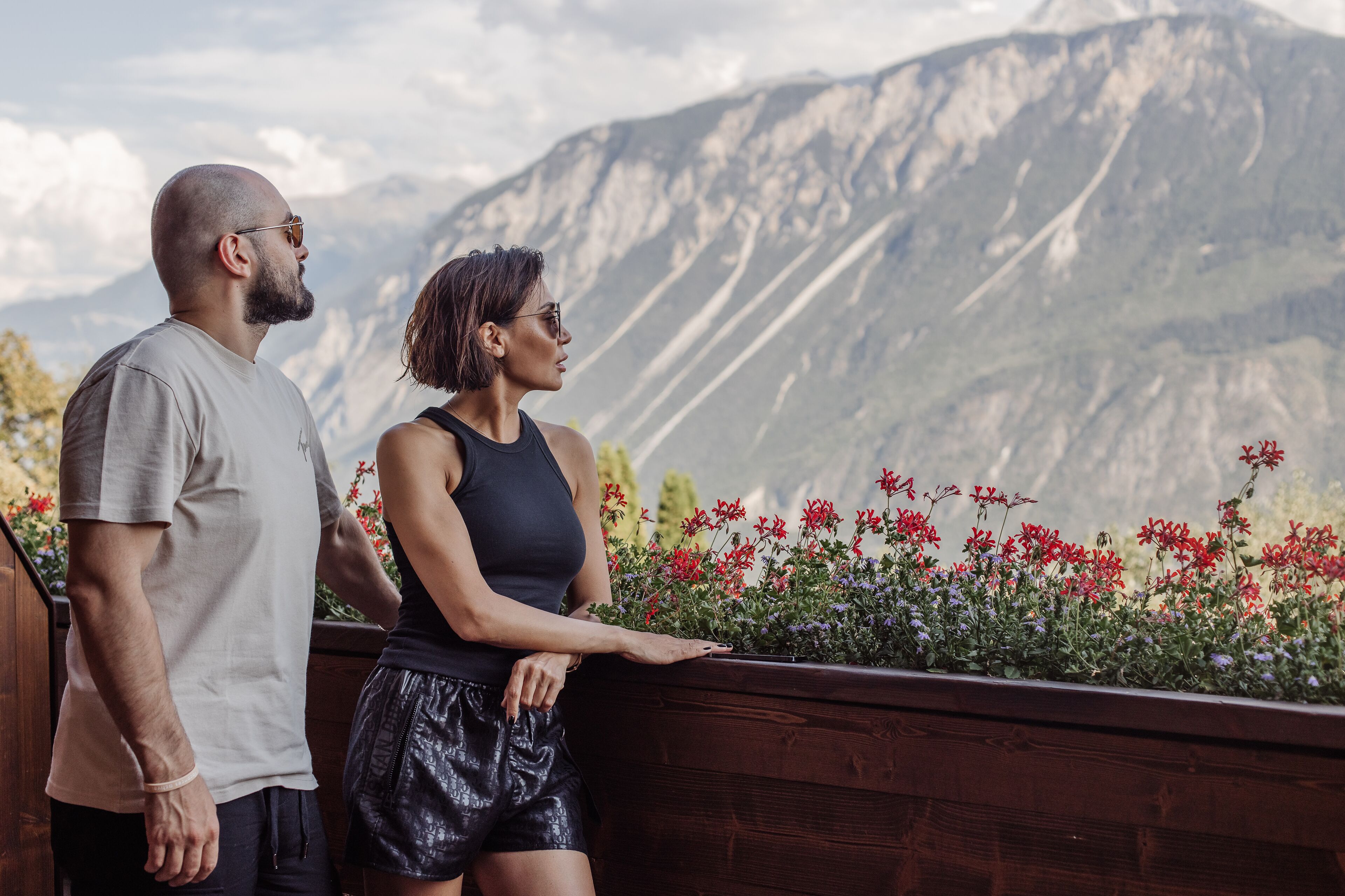 Two people standing by a wooden railing overlooking a mountainous landscape with colorful flowers in the foreground.