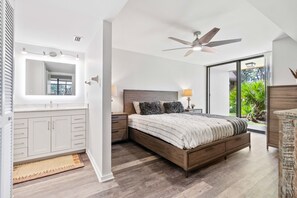 Une chambre avec un grand lit, une coiffeuse avec un miroir, un ventilateur de plafond et une vue sur la verdure à l’extérieur.