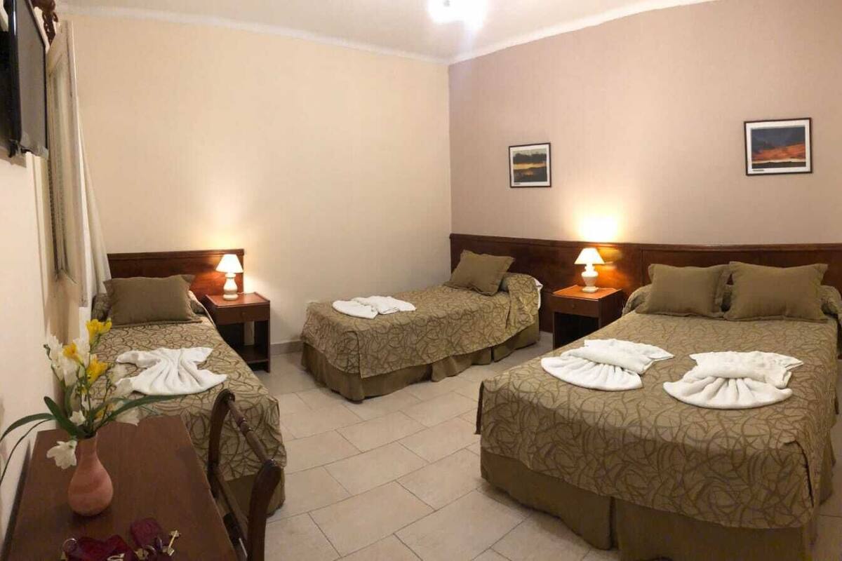 A hotel room with three beds, each with a lamp and towels. There are two framed pictures on the wall.
