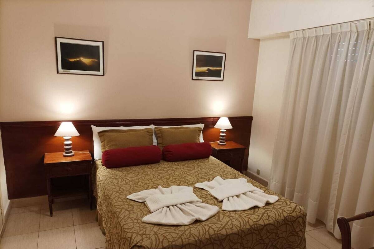A hotel room with a bed, two bedside lamps, a headboard, two framed pictures on the wall, and a window with curtains.