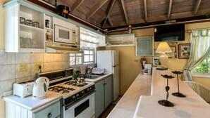 Una cocina con estufa, microondas y refrigerador. Hay una ventana con cortinas, un fregadero y una encimera con tetera y tostadora.