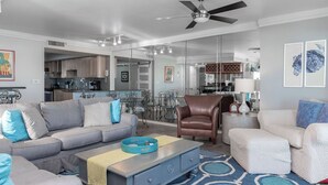 A modern living room with a grey sofa, a brown leather chair, and a white armchair. There is a coffee table with a tray and a blue rug. The room has a ceiling fan and a kitchen in the background. - BOOK NOW for a Great GetAway~2 Bedroom/ 3 Bath~Gulf Front!!!! (Gulf Shores)