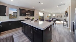 A modern kitchen with dark cabinets, a large island, and a dining area with a view. - Luxe Private Getaway in the Heart of Oceanside! (Oceanside)