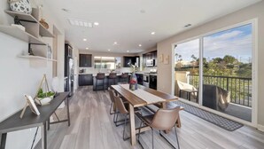 A modern kitchen with dark cabinets and a large island, a dining area with a wooden table and chairs, and a balcony with a view of the outdoors. - Luxe Private Getaway in the Heart of Oceanside! (Oceanside)